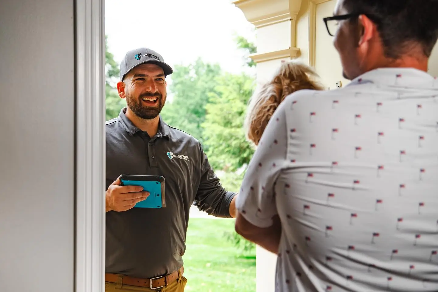 Smiling technician in branded polo and cap holding tablet, greeting homeowners at front door