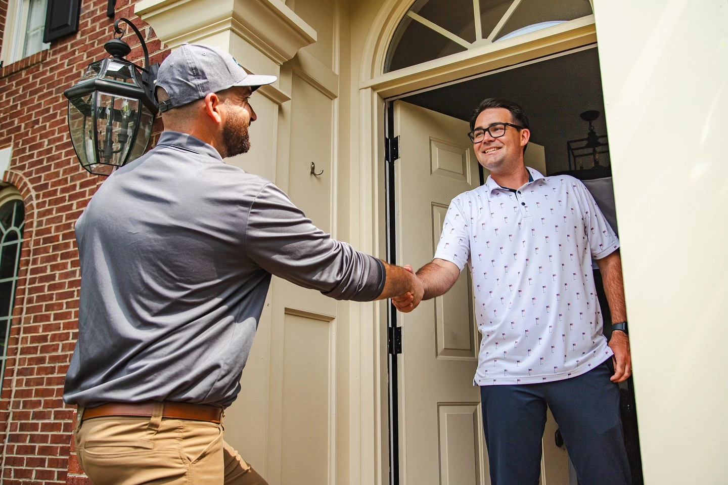 Two men shaking hands at a residential front door, service worker in gray shirt and cap meeting homeowner in light blue